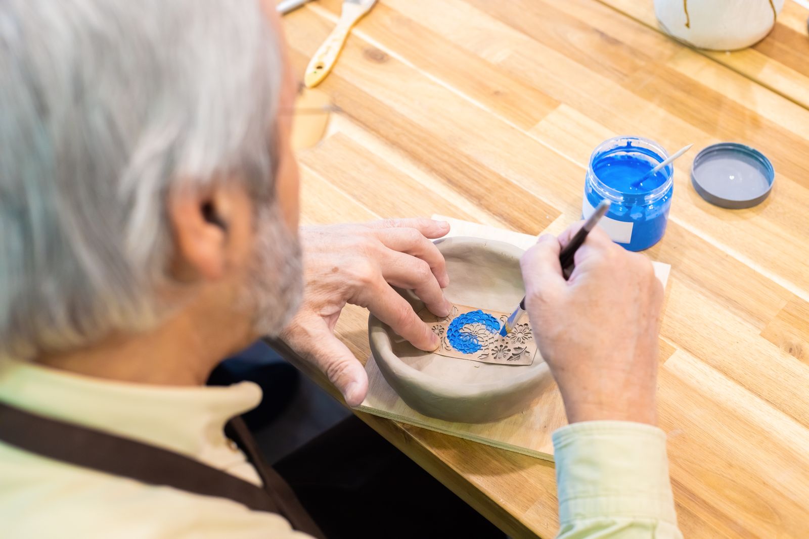 A senior man carefully painting a ceramic bowl with blue paint